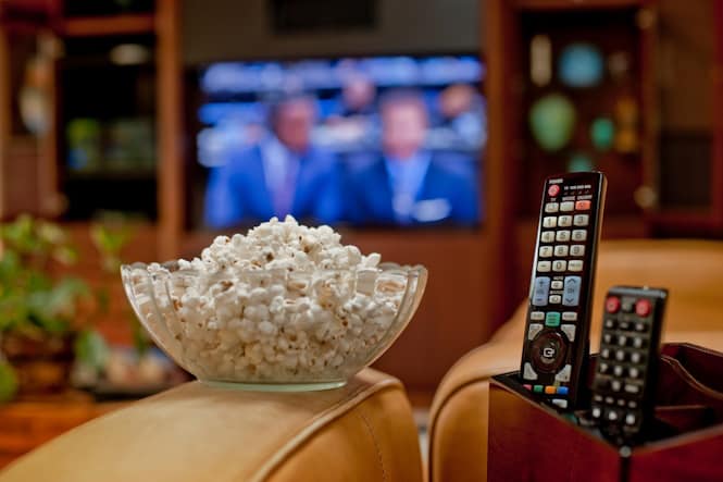 bowl of popcorn and tv remotes on furniture armrests with tv on in the background